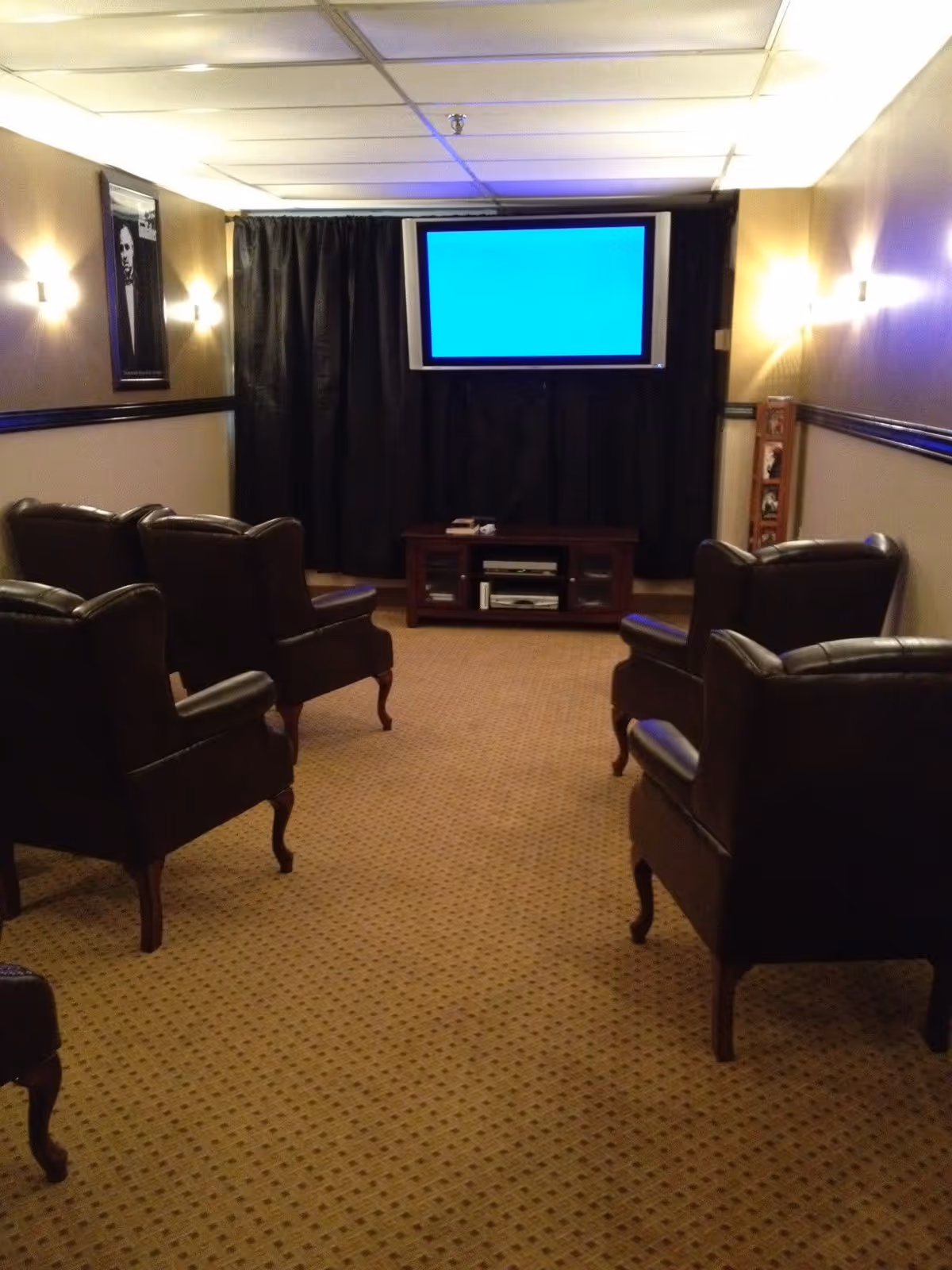 A small theater room with six dark leather armchairs arranged in two rows facing a wall-mounted flat screen TV. The room has beige walls with black trim, wall sconces providing warm lighting, a carpeted floor, and a framed picture on the left wall.