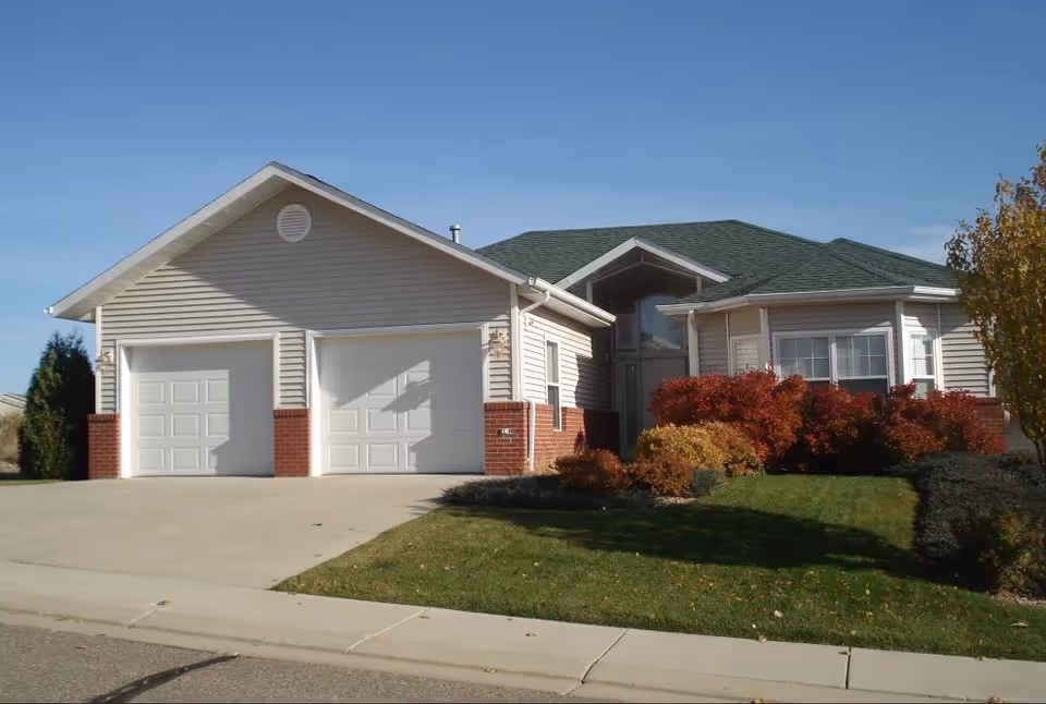 Front view of a single-story suburban building with a two-car garage, green roof and landscaped lawn with shrubs.