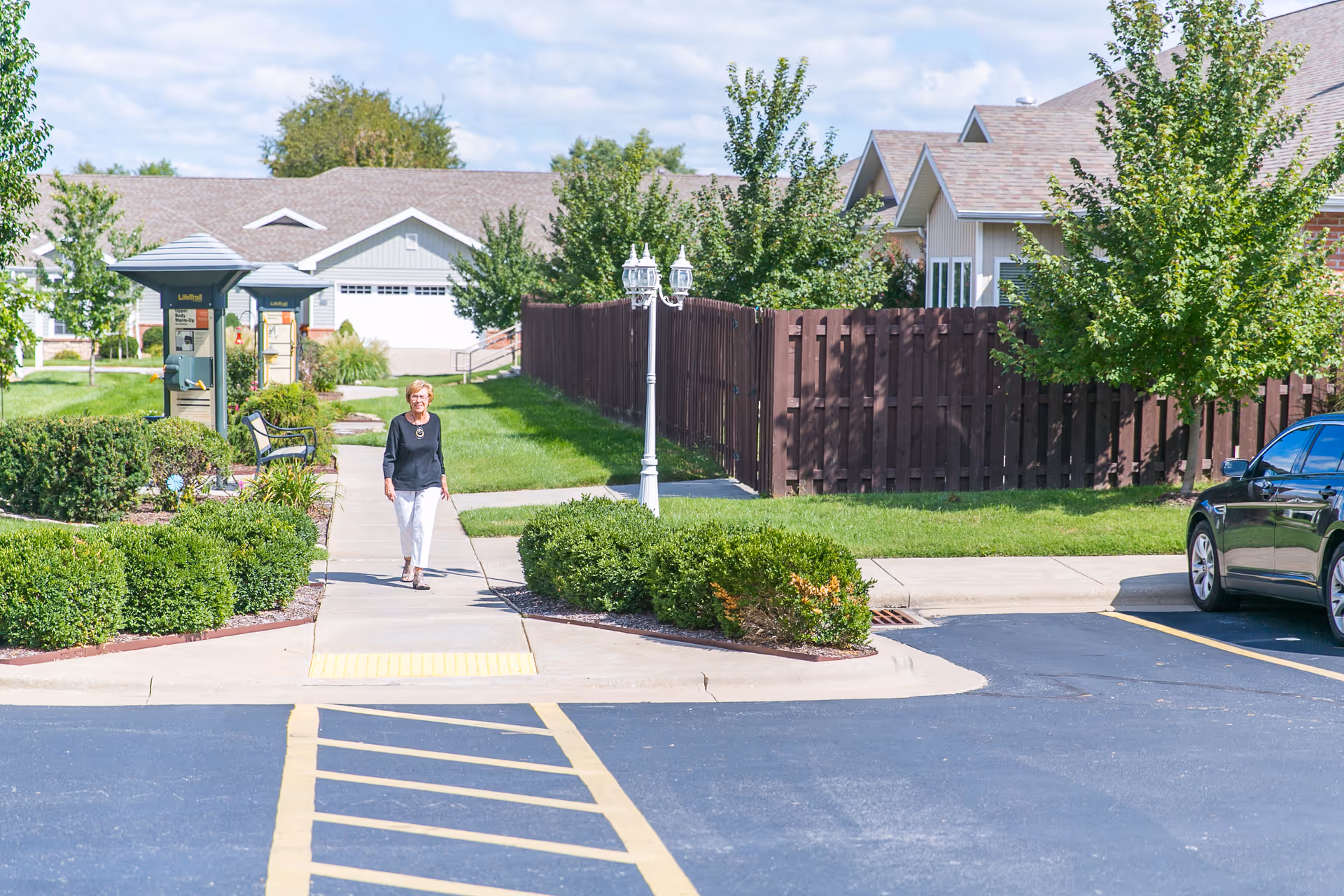 An elderly woman walking on a sidewalk in a senior living community with green bushes, trees, and residential buildings in the background on a sunny day.
