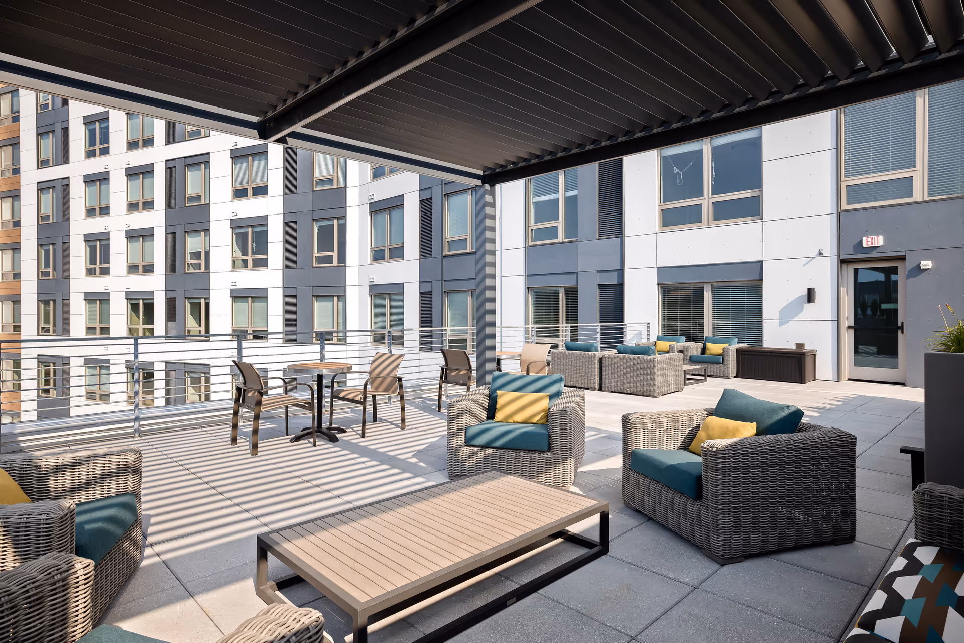 Outdoor patio area at Revelle King of Prussia with modern wicker seating including armchairs and sofas with blue and yellow cushions, a wooden coffee table, and a small round table with chairs. The space is shaded by a slatted pergola and surrounded by a metal railing, with the exterior of a multi-story building visible in the background.