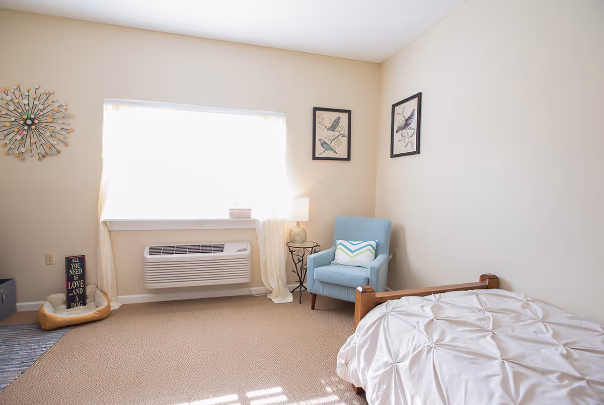 Bright single-occupancy bedroom with a bed, blue armchair, window air conditioner and framed wall art.