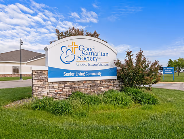 Outdoor view of a large stone and white sign for Good Samaritan Society Grand Island Village Senior Living Community, with a building and green lawn in the background under a partly cloudy blue sky.