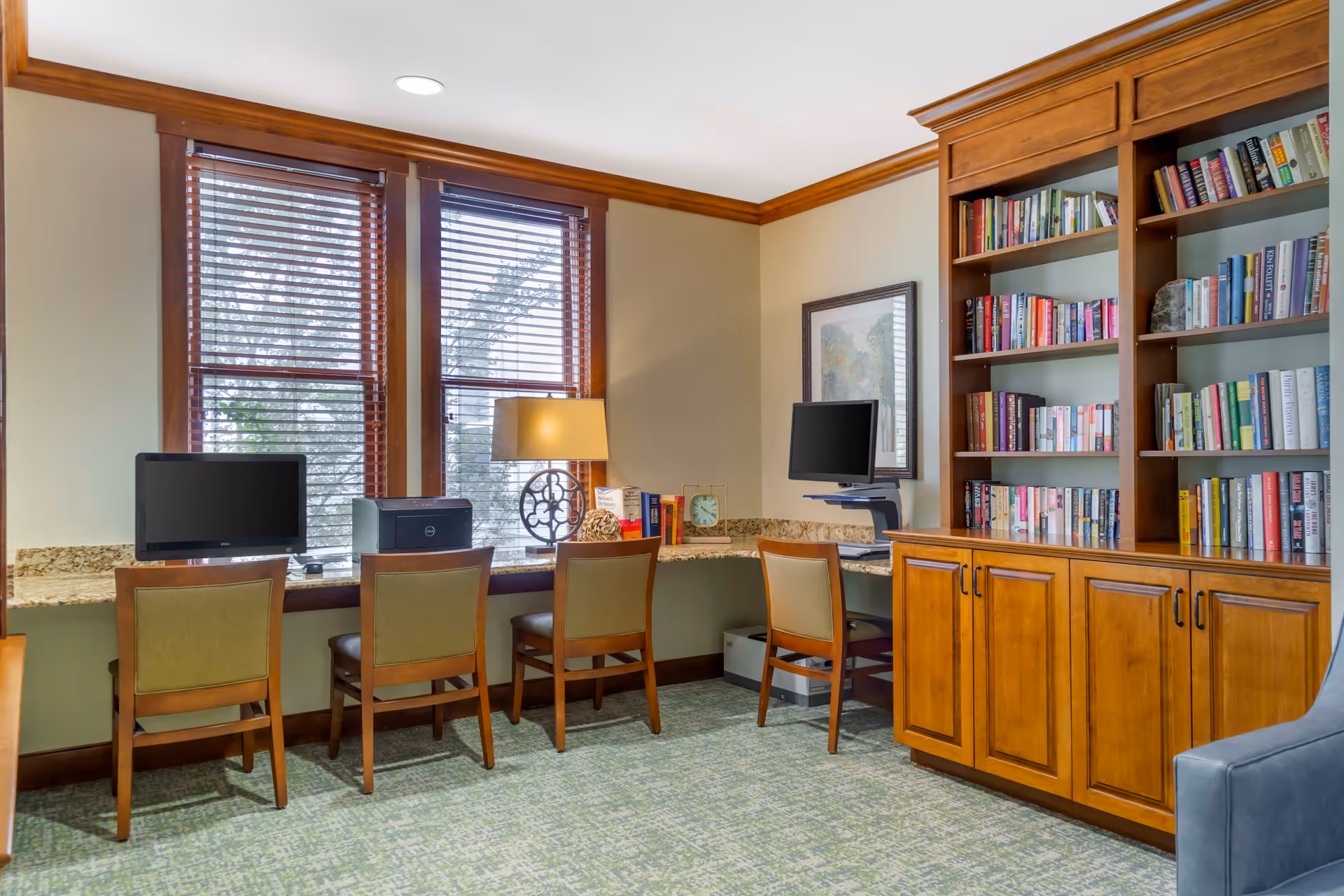 A cozy room with a long granite countertop along the wall under two large windows with wooden blinds. Four wooden chairs with green cushions are placed in front of the countertop, which holds two computer monitors, a printer, a decorative lamp, and some books. To the right, there is a wooden bookshelf filled with books and cabinets below. The room has beige walls, wooden trim, and a green patterned carpet.