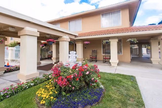 Front entrance courtyard of a beige retirement building with columns, rocking chairs, hanging flower baskets, and a central flower bed with a small statue fountain.