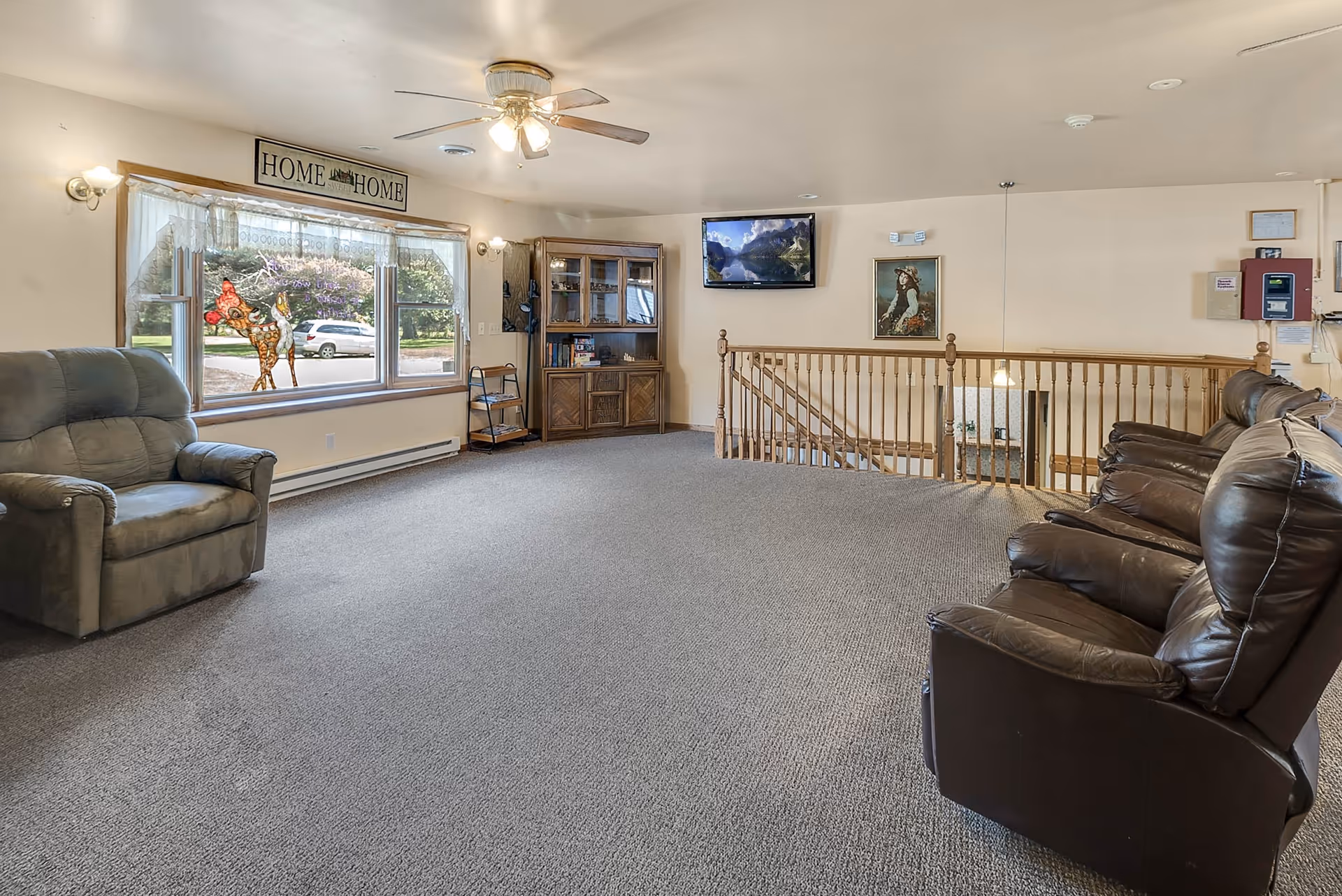 A spacious living room area with carpeted floor, featuring a large window with sheer curtains and a decorative sign above it that reads 'Home Sweet Home'. There are two armchairs on the left and three leather recliners on the right. A wooden cabinet with glass doors and shelves is against the far wall, with a flat-screen TV mounted above it. A wooden railing separates the living area from a staircase leading downstairs. The walls are light-colored, and the ceiling has a ceiling fan with lights.