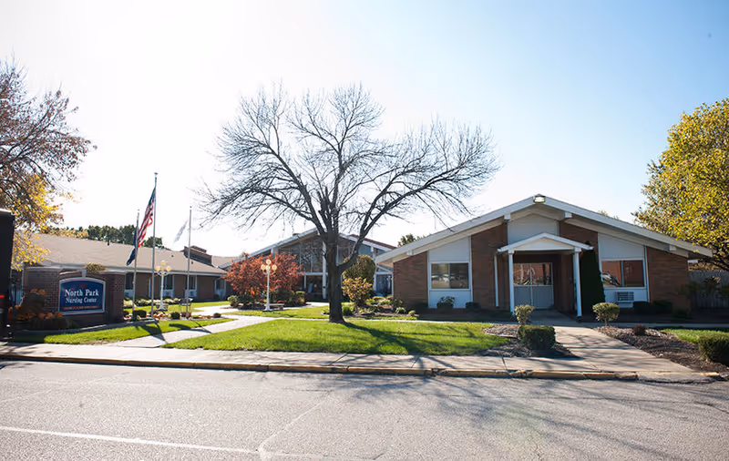 Front view of North Park Nursing Center: a single-story brick building with a central entrance, lawn, trees, flagpoles, and a sign.