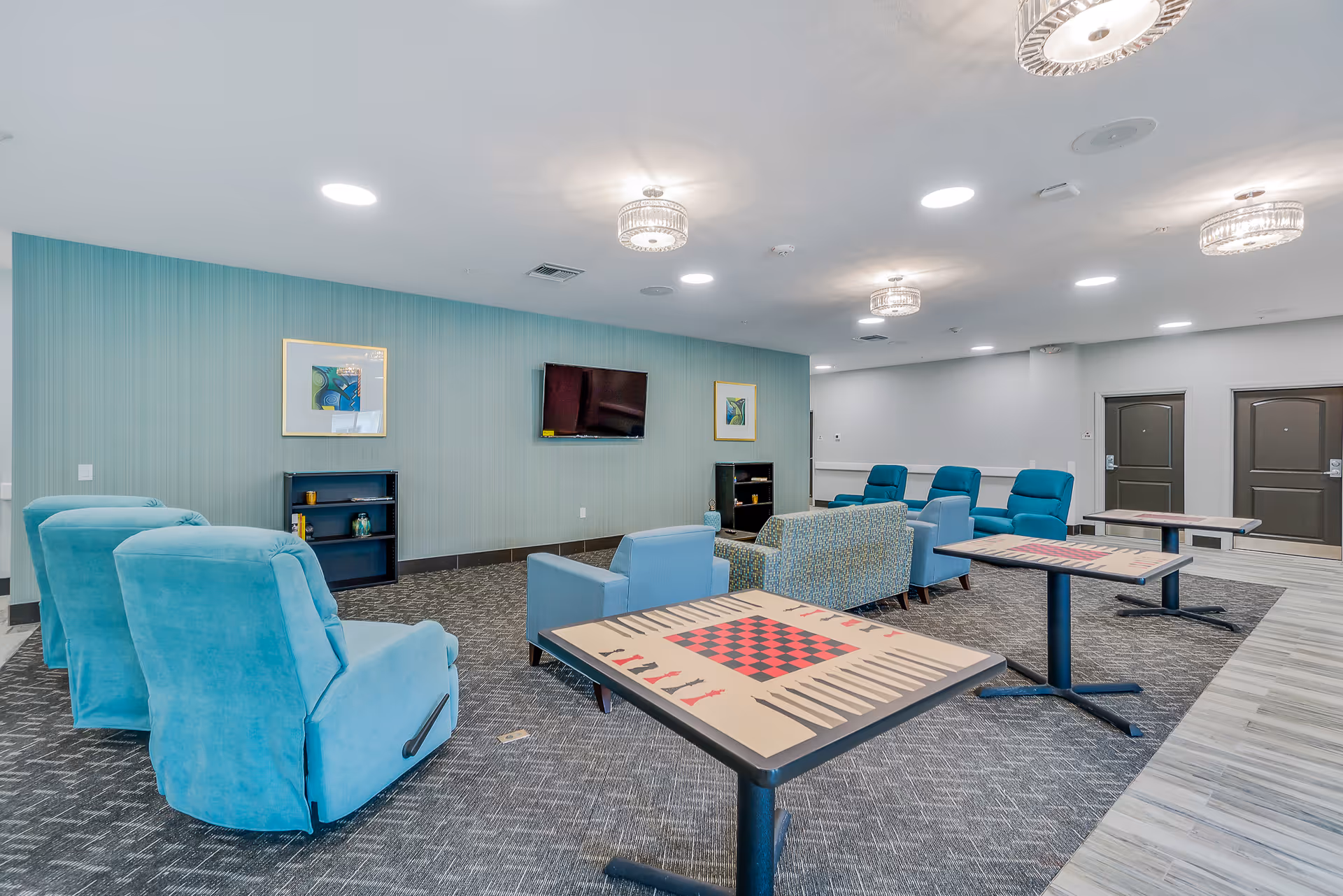 A spacious common area in Senior Point Assisted Living featuring multiple blue upholstered armchairs and sofas arranged around game tables with checkerboard and backgammon patterns. The room has a teal accent wall with a mounted flat-screen TV and framed artwork, modern ceiling lights, and a carpeted floor with a tiled walkway.