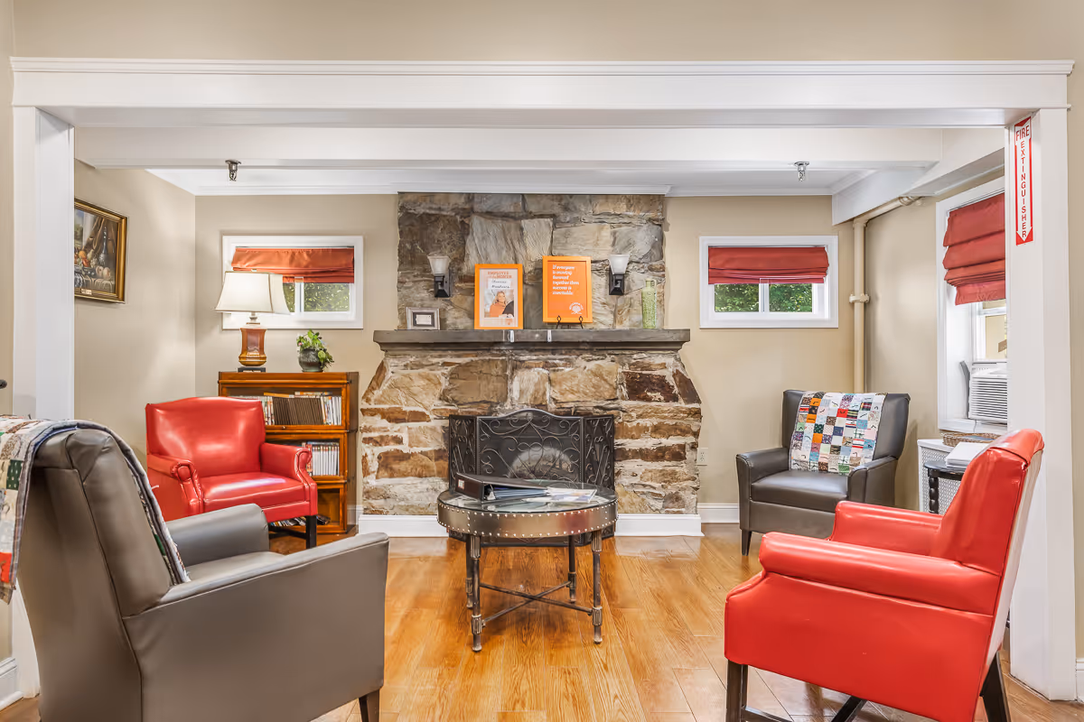 A cozy living room area with a stone fireplace in the center, flanked by two small windows with red Roman shades. Four armchairs, two red and two gray, are arranged around a round metal coffee table. A wooden bookshelf with a lamp and some decorations is on the left side. The room has wooden flooring and light-colored walls.