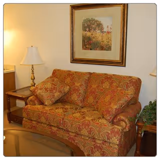 A cozy living room area featuring a patterned red and gold sofa with matching cushions, a wooden side table with a brass lamp, a framed floral painting on the wall, and a glass coffee table in the foreground.