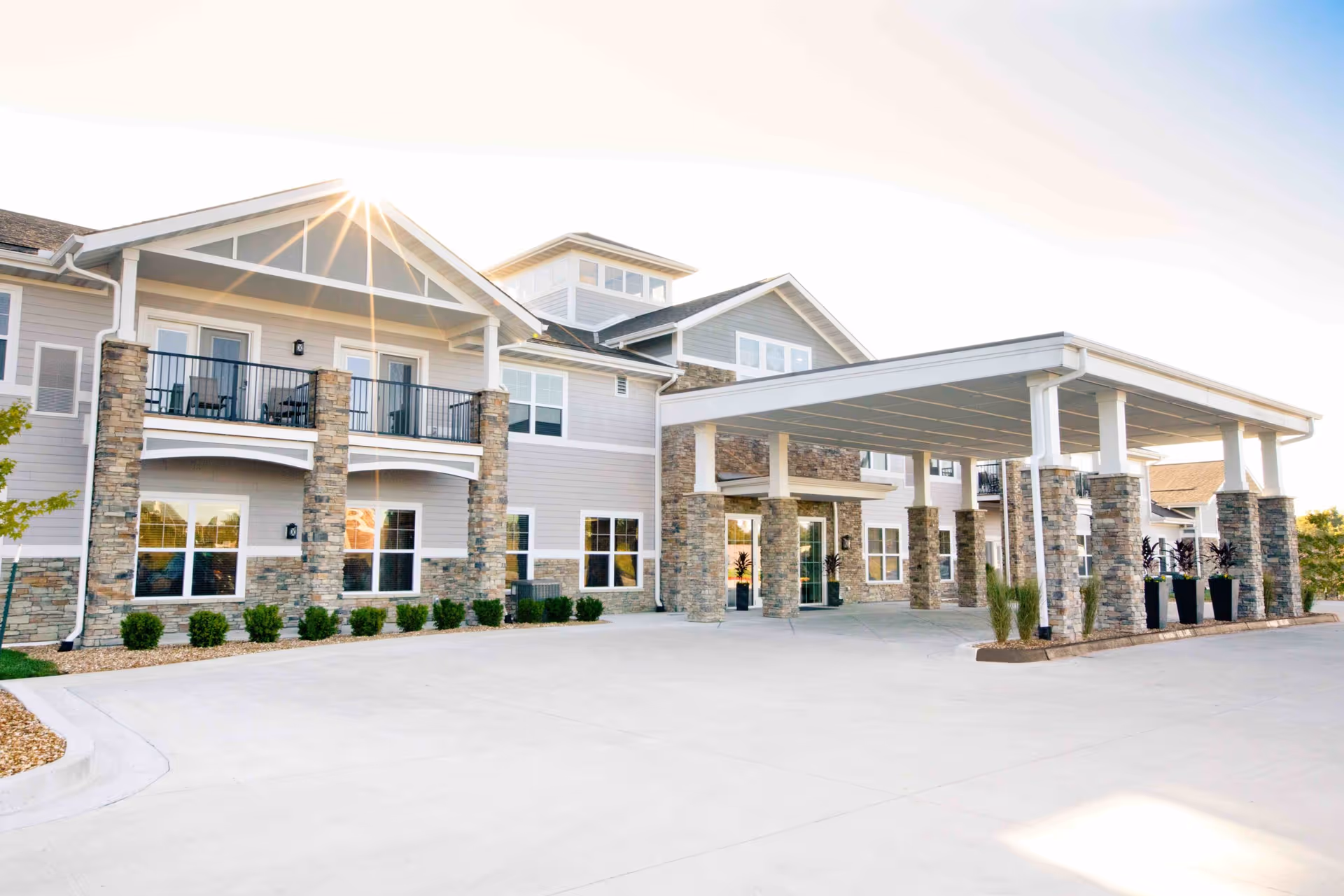 Exterior front view of Cedarhurst Senior Living of Blue Springs building with stone pillars, balconies, and a covered entrance area under a clear sky with the sun shining.