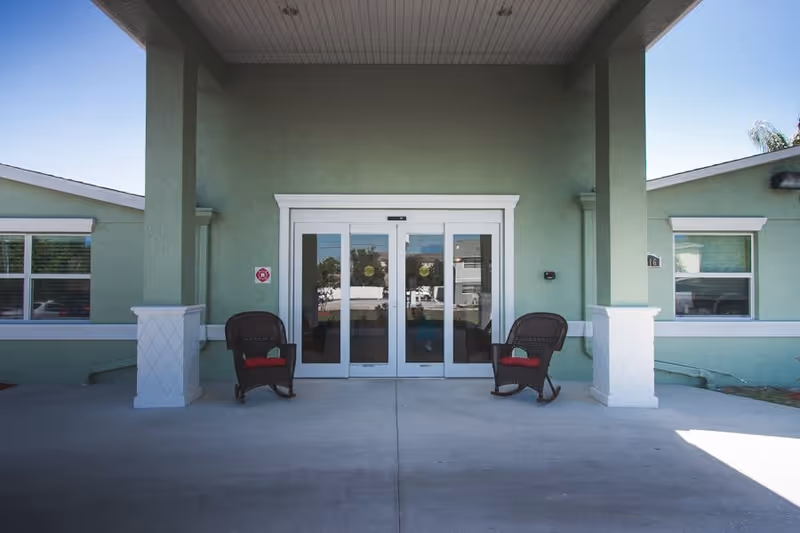 Entrance to a building with green walls and white trim, featuring double glass sliding doors. Two dark wicker rocking chairs with red cushions are placed on either side of the entrance under a covered porch.