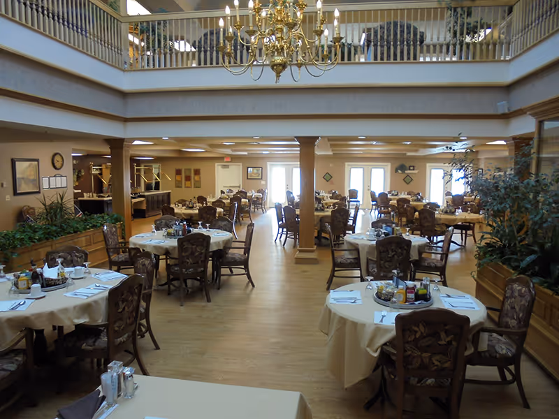 Spacious dining room in an assisted living community with multiple round tables covered with beige tablecloths, each set with chairs, napkins, condiments, and place settings. The room features wooden flooring, large windows letting in natural light, indoor plants, and a decorative chandelier hanging from the ceiling.