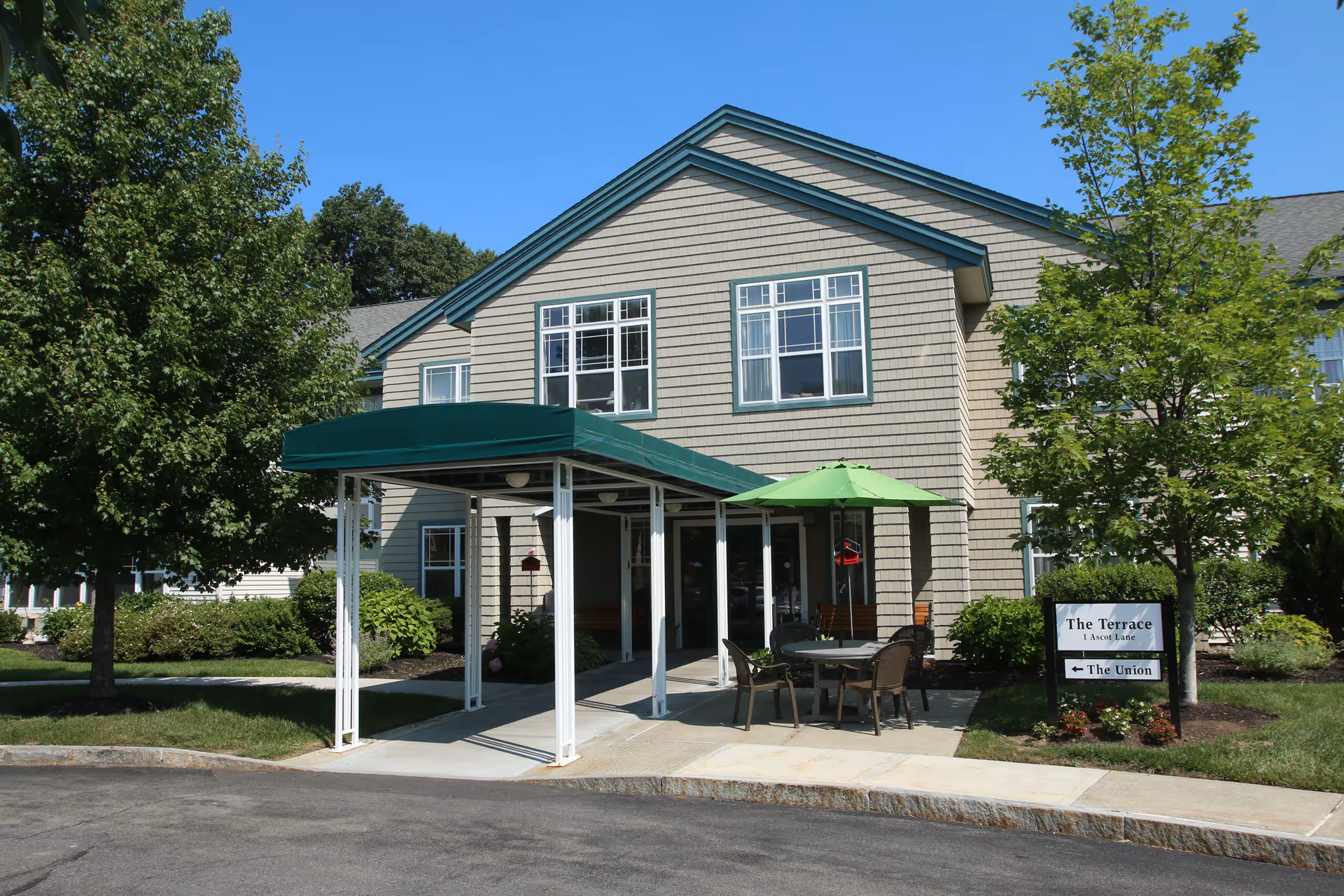 Exterior view of a senior living facility building with beige siding and green trim. There is a covered entrance with a green canopy, a patio area with a table, chairs, and a green umbrella. Trees and shrubs surround the building, and a sign near the entrance reads 'The Terrace 1 Ascot Lane' with an arrow pointing to 'The Union'.