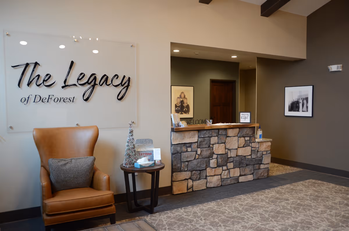 Reception area of The Legacy of DeForest featuring a stone front desk with a welcome sign, a brown leather armchair with a gray pillow, a small round table with a decorative Christmas tree and tissues, and wall art including a large sign with the facility name and framed photographs.