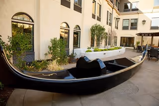 Outdoor courtyard with a black gondola-style bench on a paved patio beside planter beds and arched building windows.
