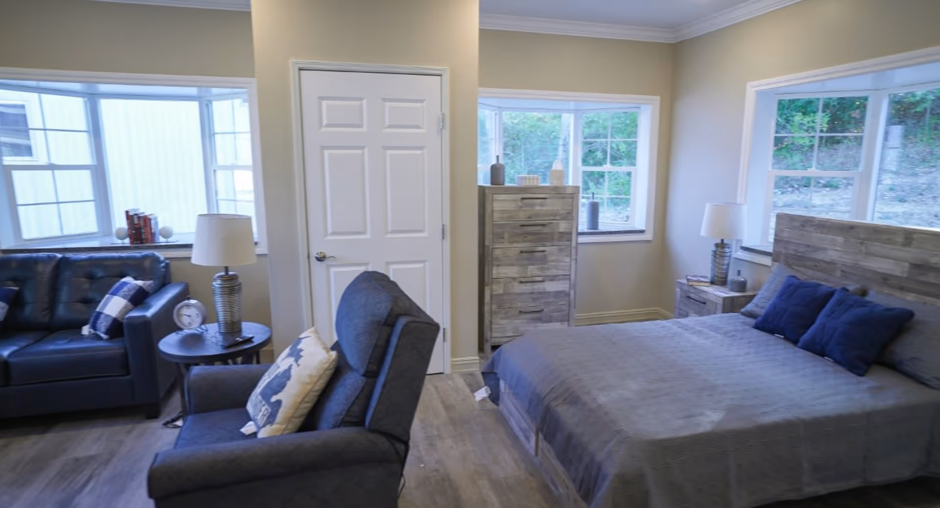 Bright studio-style bedroom with a bed, dresser, armchair and sofa near multiple windows.