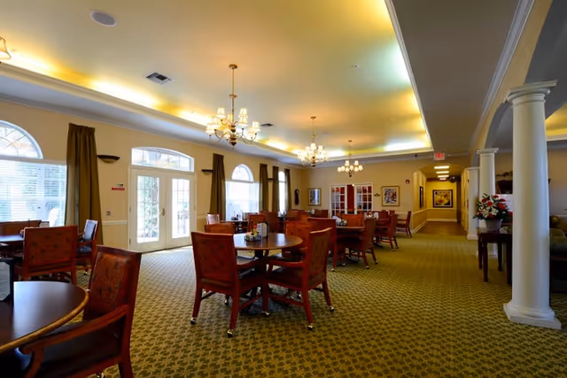 A spacious dining room with multiple round wooden tables and cushioned chairs arranged neatly. The room features large windows with green curtains, chandeliers hanging from the ceiling, and a patterned green carpet. There are columns on the right side and framed artwork on the walls.