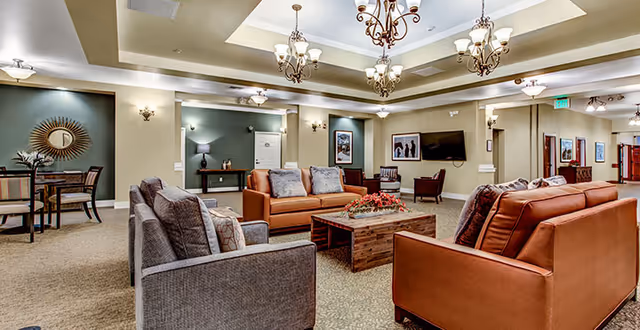 A spacious and well-lit living room area in an assisted living facility featuring comfortable seating including two brown leather sofas and two gray armchairs arranged around a wooden coffee table with a floral centerpiece. The room has beige walls, carpeted floors, decorative wall sconces, chandeliers, and framed artwork on the walls. In the background, there is a small dining area with a round table and chairs, and a flat-screen TV mounted on the wall.