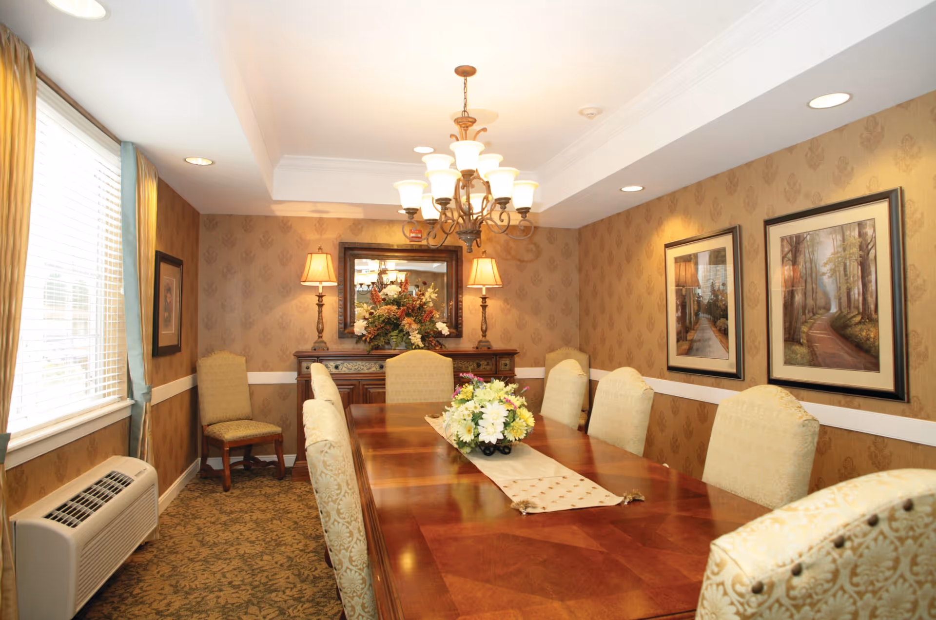 A warmly lit dining room with a long wooden table surrounded by upholstered chairs. The table has a decorative runner and a floral centerpiece. The walls are adorned with patterned wallpaper and framed landscape paintings. Two table lamps and a chandelier provide lighting, and a window with curtains lets in natural light.
