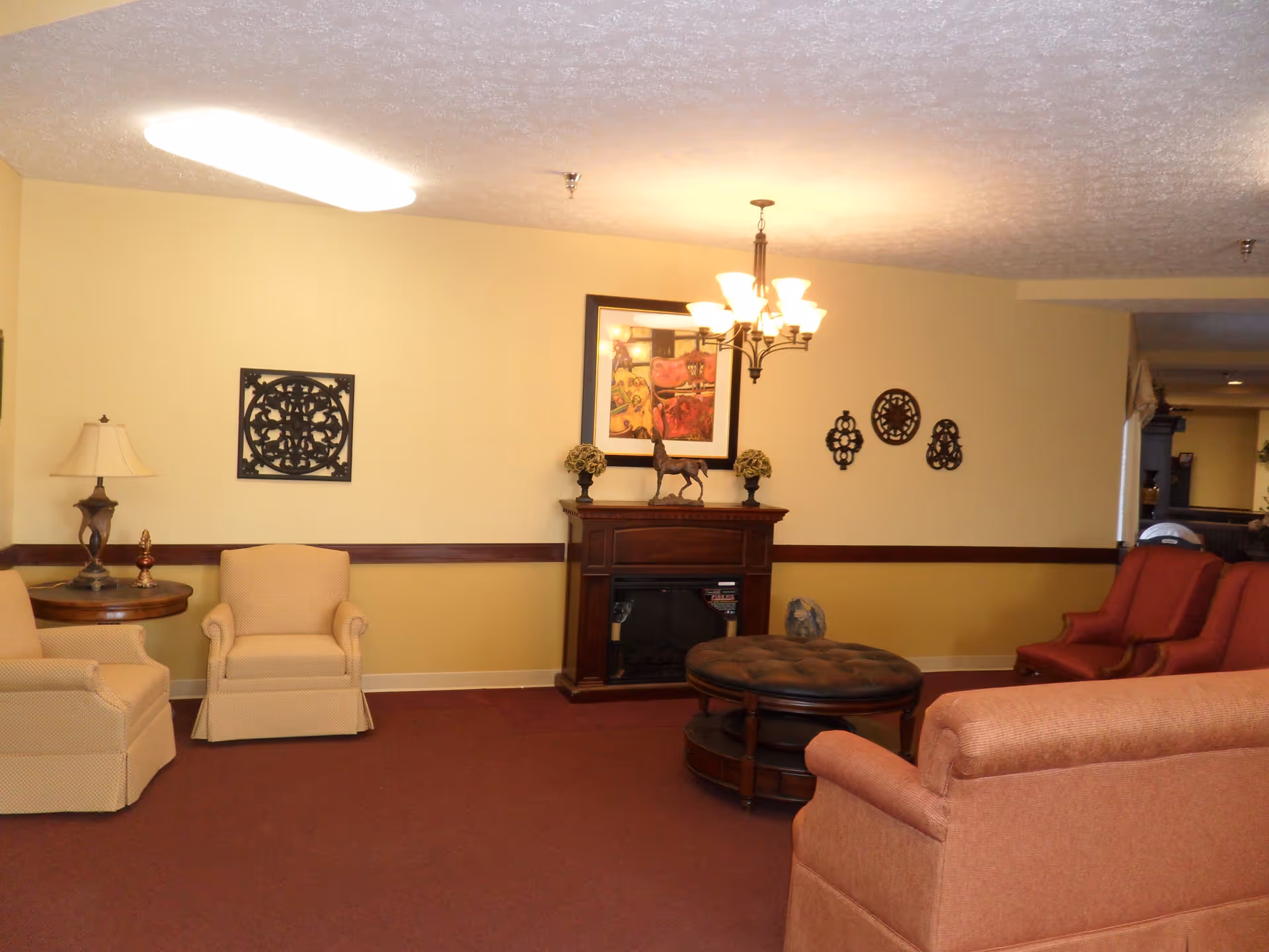 A cozy living room area with beige and red upholstered chairs, a round wooden coffee table with a cushioned top, a wooden fireplace mantel decorated with a horse statue and two small plants, wall art, and a chandelier hanging from the ceiling.