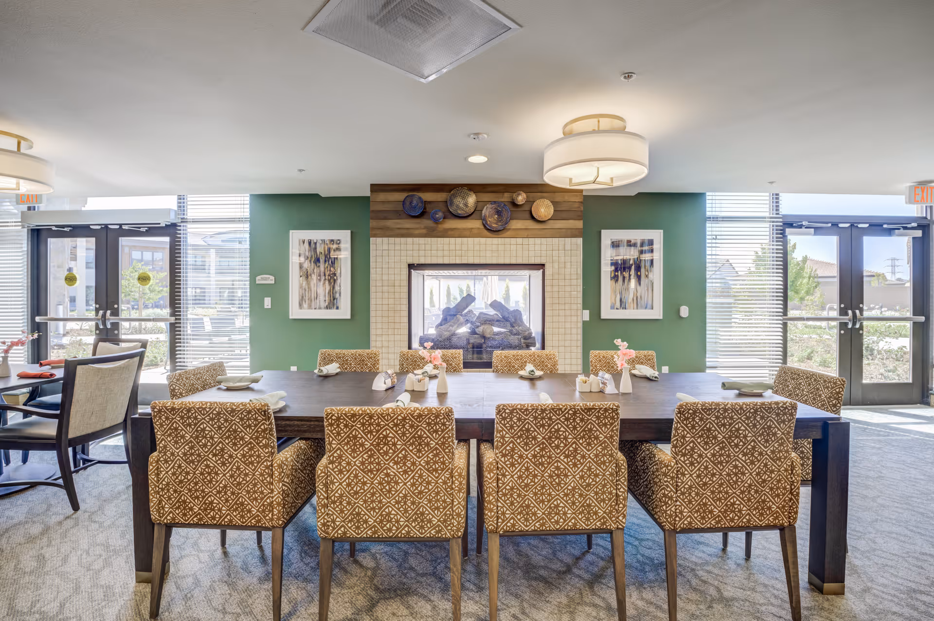 A dining area in a senior living facility with a large dark wooden table surrounded by patterned upholstered chairs. The table is set with napkins and small flower vases. Behind the table is a double-sided fireplace with a decorative wall featuring plates and two framed artworks on green walls. Large windows and glass doors on either side let in natural light.