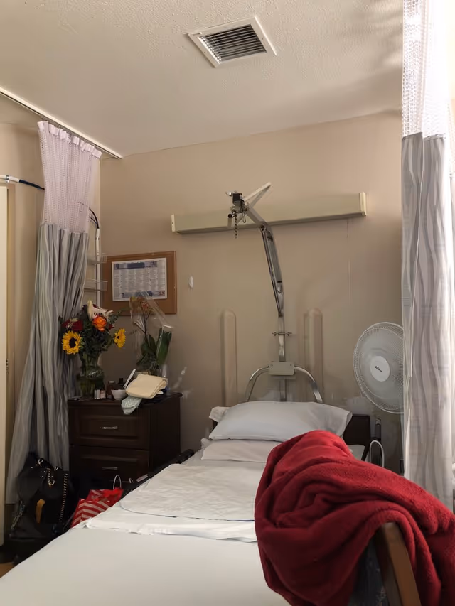 Hospital-style patient bedroom with a bed, privacy curtains, dresser, flowers, and a fan.