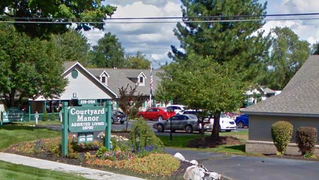 Exterior view of Courtyard Manor assisted living facility showing a green sign with the facility name, surrounded by landscaped greenery, trees, and parked cars in the background under a partly cloudy sky.