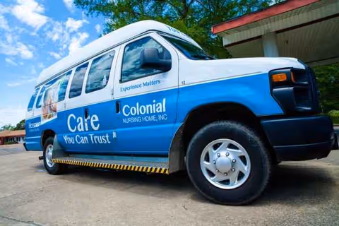 Blue and white Colonial Nursing Home transport van is parked outside a building.