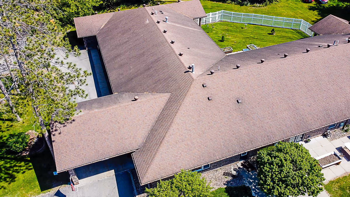 Aerial view of a senior living facility building with a brown shingled roof, surrounded by green grass, trees, and a white fenced area in the backyard.