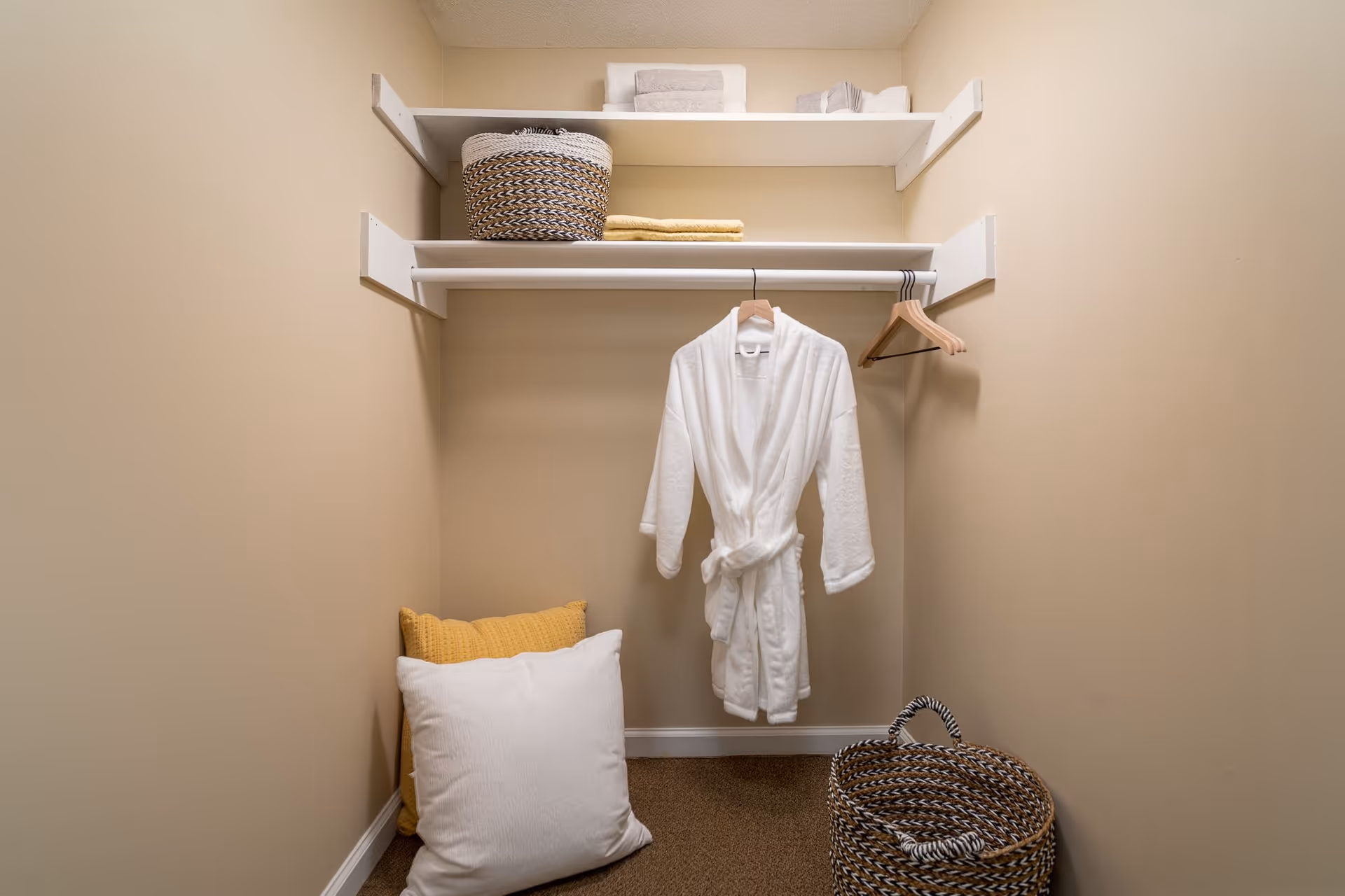 Small walk-in closet with a white bathrobe hanging on a rod, shelves holding baskets and towels, and pillows plus a woven basket on the floor.