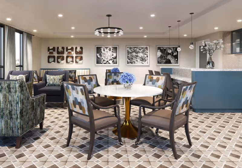 A modern and stylish common area with a round white table surrounded by six patterned chairs. The floor has a geometric tile design. In the background, there are two armchairs and a blue counter with pendant lights hanging above it. The walls are decorated with black and white framed photographs and a decorative wall piece. A vase with blue flowers is placed on the table, and another vase with white flowers is on the counter.
