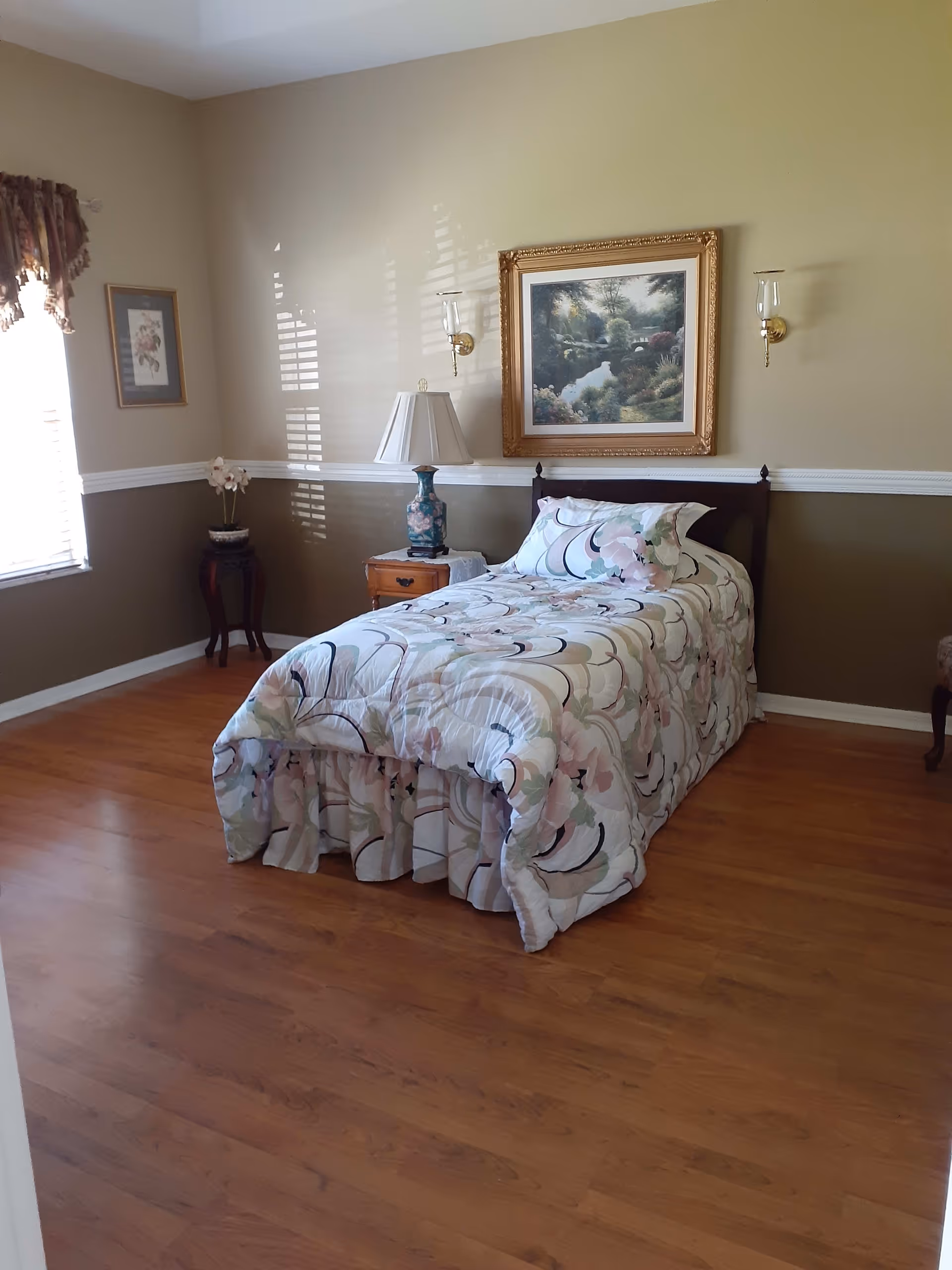 A neatly made single bed with floral patterned bedding in a bedroom with wooden flooring. The room has beige walls with a white chair rail, a framed landscape painting above the bed, a bedside table with a decorative lamp, a small plant stand with a flower pot, and a window with a curtain letting in natural light.