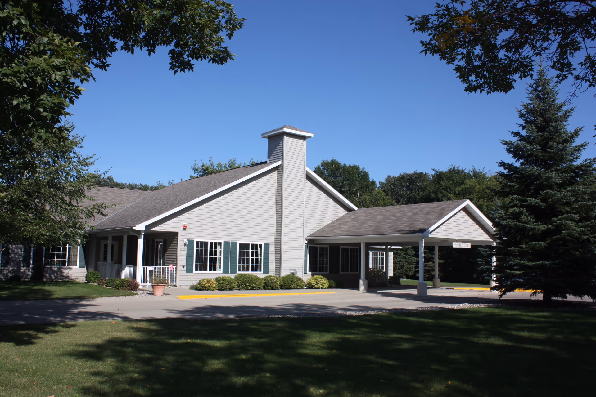 Exterior view of a single-story building with beige siding and green shutters, surrounded by trees and a well-maintained lawn under a clear blue sky. The building has a covered entrance driveway and a small porch area.