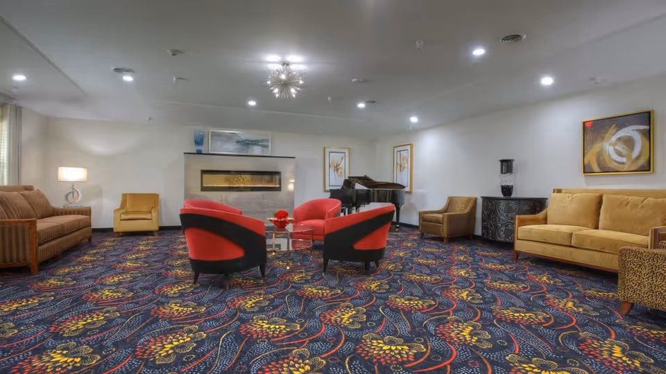 Spacious lounge with colorful patterned carpet, red armchairs around a glass table, sofas, a fireplace and a grand piano.