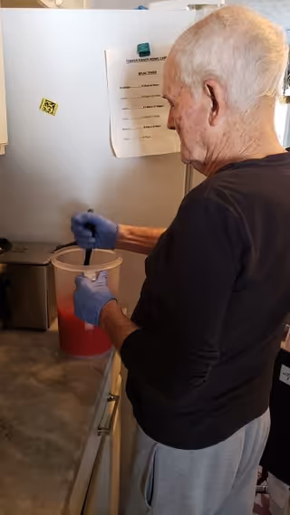 An elderly person wearing blue gloves is stirring a red liquid in a large plastic container in a kitchen. A meal menu from Tender Hands Home Care is taped to the refrigerator in the background.