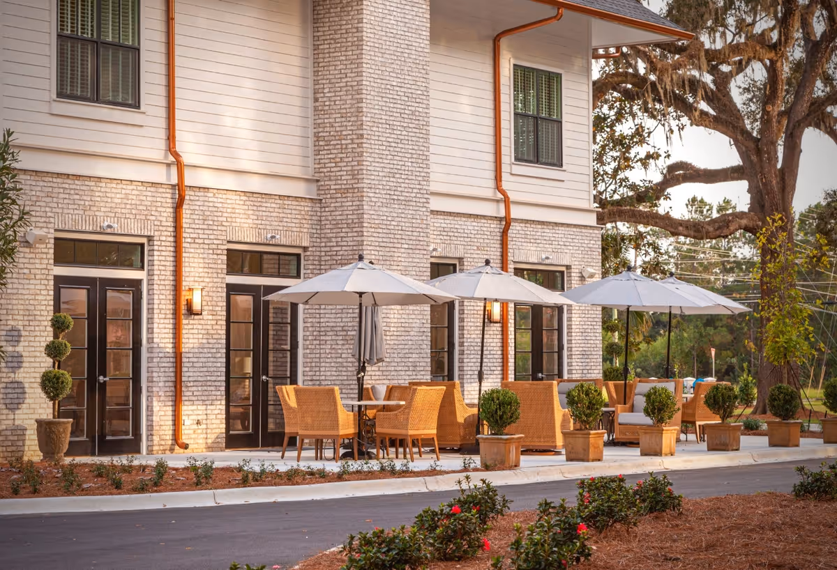 Outdoor patio area of a senior living facility with wicker chairs and tables under large white umbrellas. The building has light brick walls, black-framed glass doors, and copper downspouts. There are potted plants and small bushes lining the patio, with a large tree and greenery in the background.