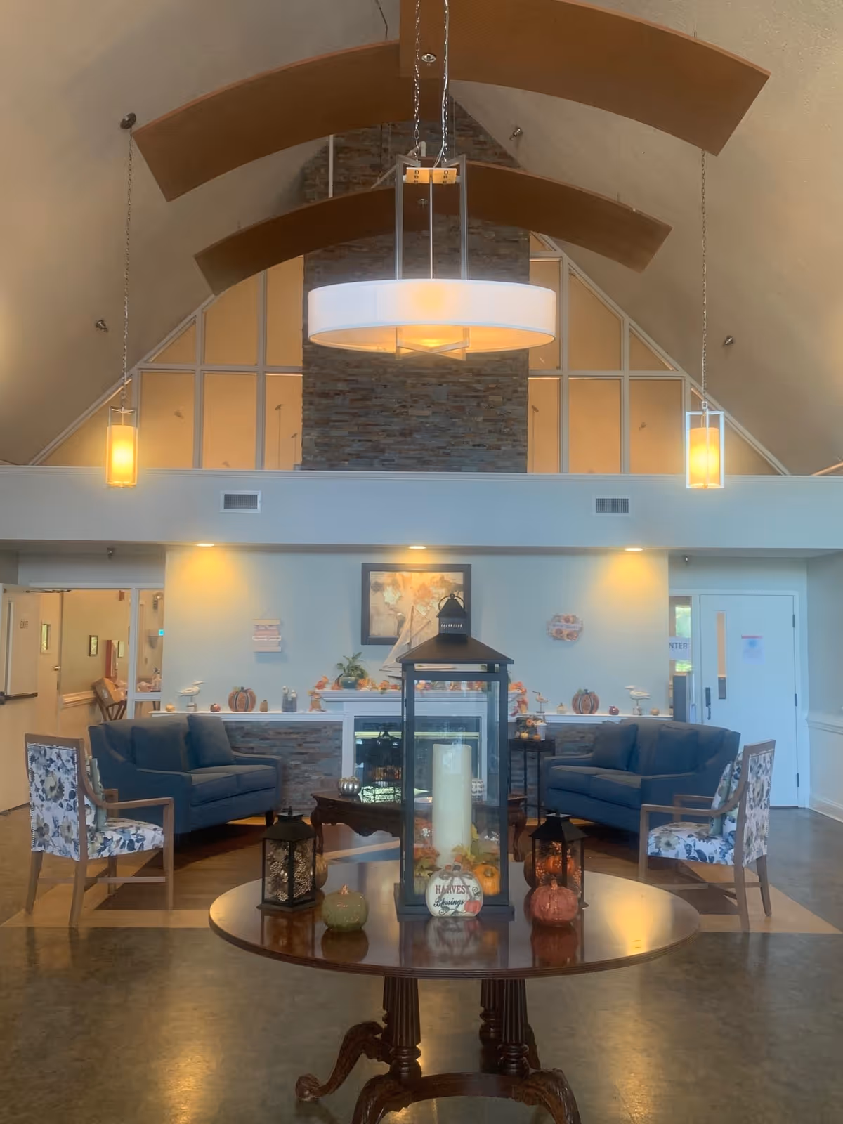 A cozy living room area with two blue sofas and two floral armchairs arranged around a fireplace. A round wooden table in the foreground holds decorative lanterns and small pumpkins. The room features a high vaulted ceiling with wooden beams and modern hanging light fixtures. The walls are light-colored, and there are large windows above the fireplace.