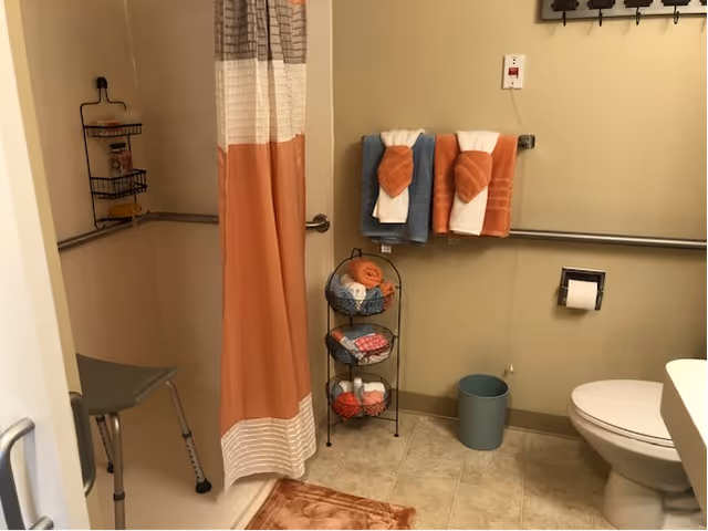 A bathroom with a shower area featuring a shower chair and a curtain in shades of orange and white. There are towels hanging on a rail and neatly rolled towels on a three-tier metal rack. A toilet, a small trash bin, and a toilet paper holder are also visible. The walls are beige, and the floor is tiled.