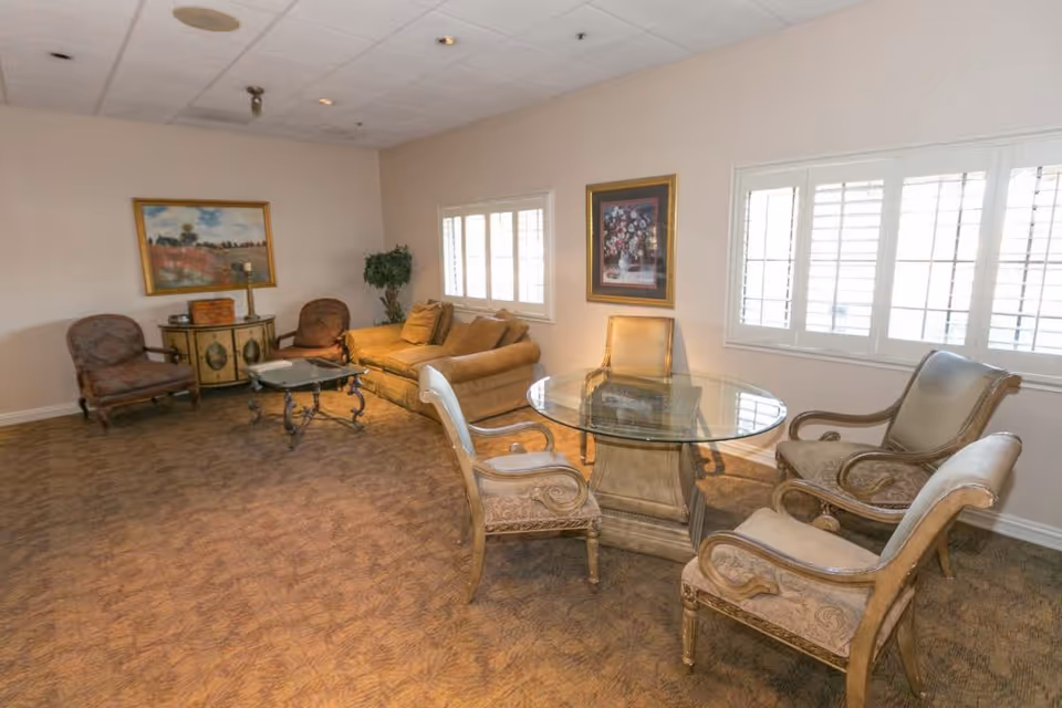 A cozy living room with a beige sofa, two upholstered armchairs, a glass-top coffee table, and a round glass dining table with four ornate chairs. The room has carpeted flooring, two framed paintings on the walls, and windows with white shutters letting in natural light.