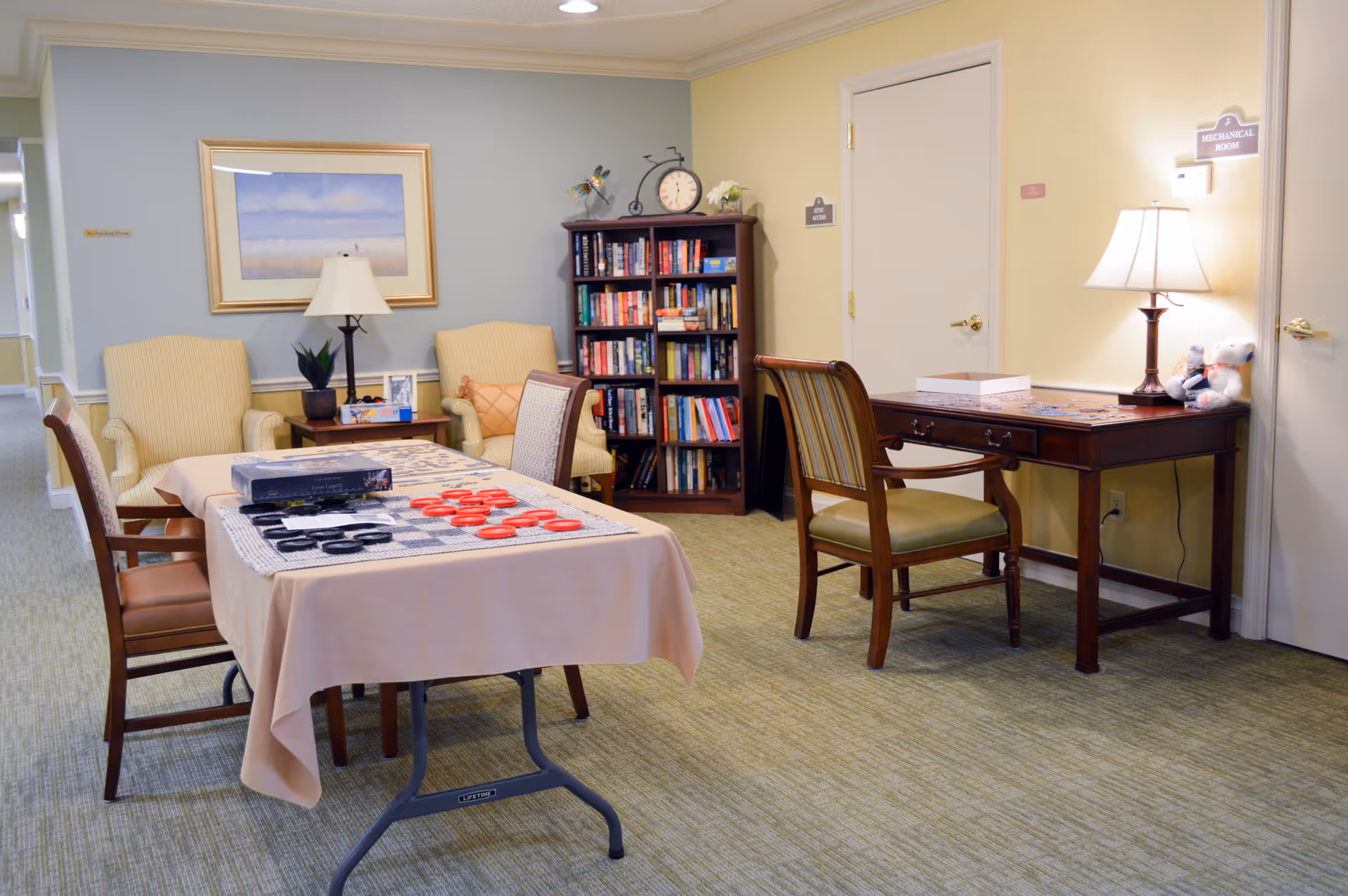 Well-lit common room with a table set up for checkers, chairs, a bookshelf, and a desk with a lamp.
