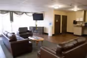 A common area in a senior living facility featuring brown leather sofas arranged around a wooden coffee table, a wall-mounted flat-screen TV, a water dispenser, and a kitchenette area with cabinets and a countertop. The room has large windows with sheer curtains allowing natural light to enter.