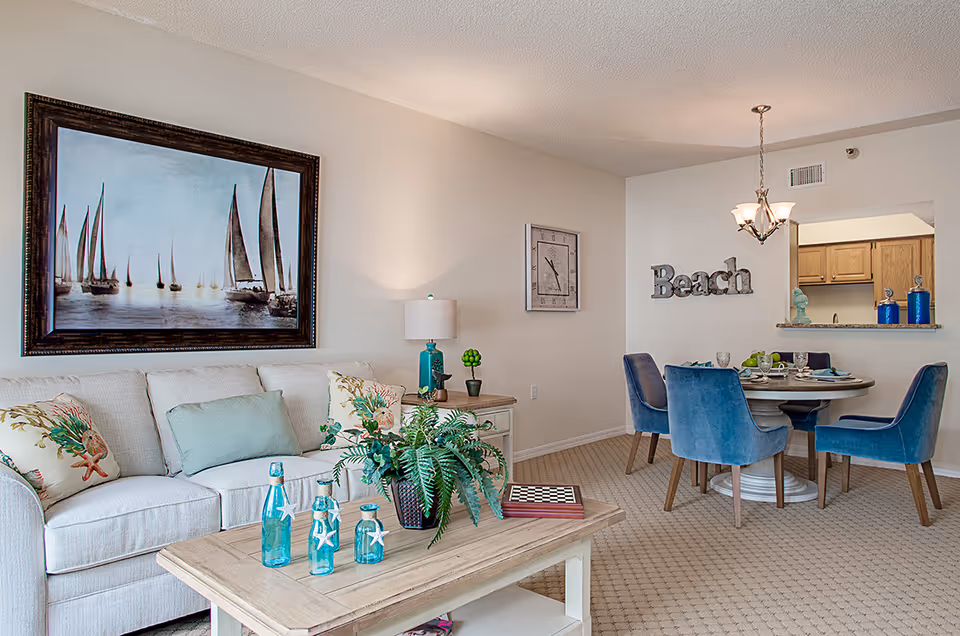 Bright coastal-styled living area with a white sofa, coffee table with blue glass bottles and plants, and a dining table with blue chairs under a chandelier.