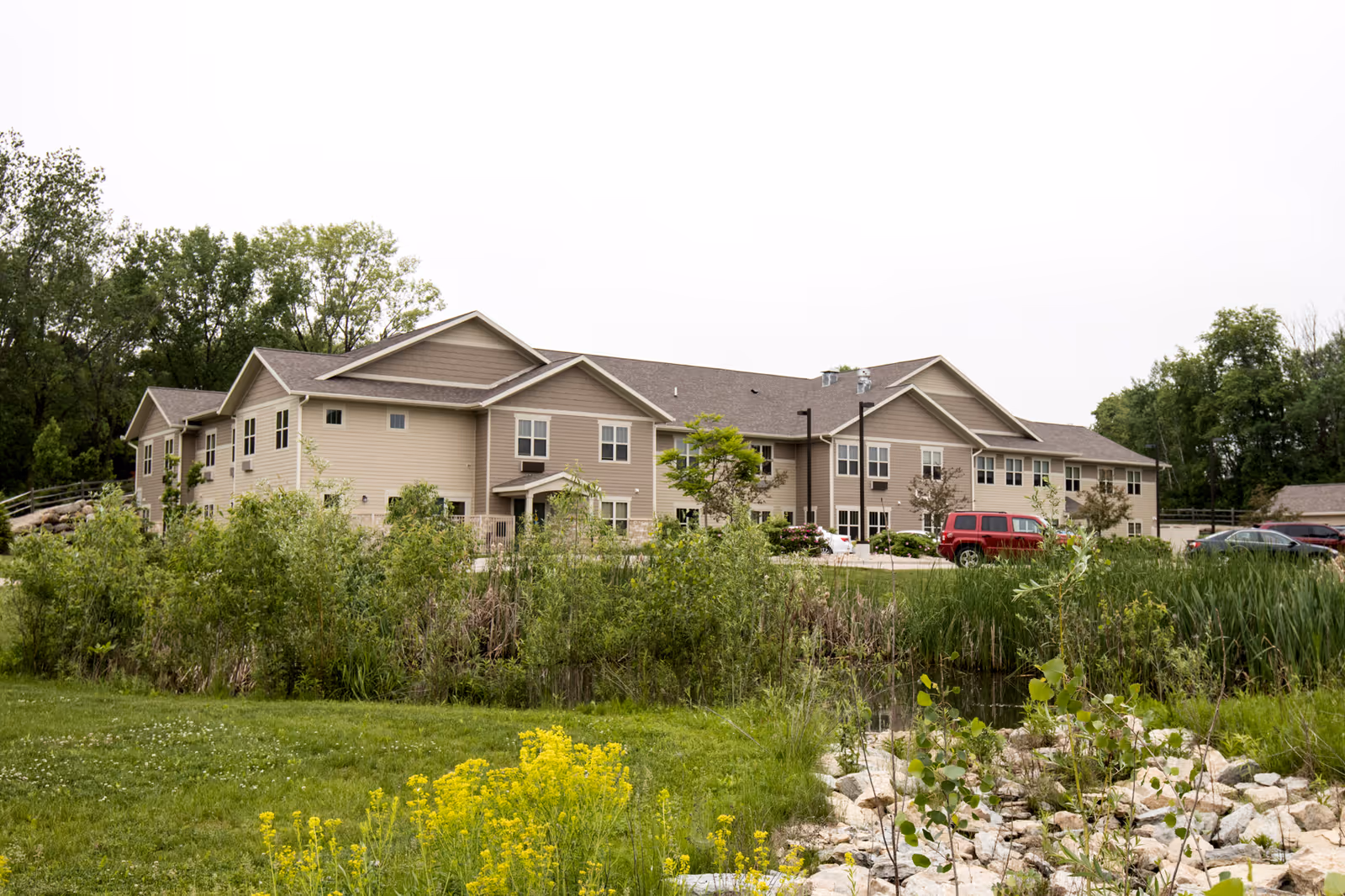 Exterior view of a two-story beige assisted living facility building surrounded by greenery, trees, and a small pond with rocks in the foreground. Several cars are parked near the building under an overcast sky.