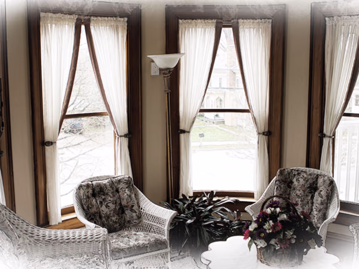 A cozy sitting area with three white wicker chairs featuring floral cushions arranged around a white table with a basket of colorful flowers. The room has large windows with sheer white curtains tied back, allowing natural light to brighten the space. A tall floor lamp stands between two of the windows, and a green plant is placed near the windowsill.