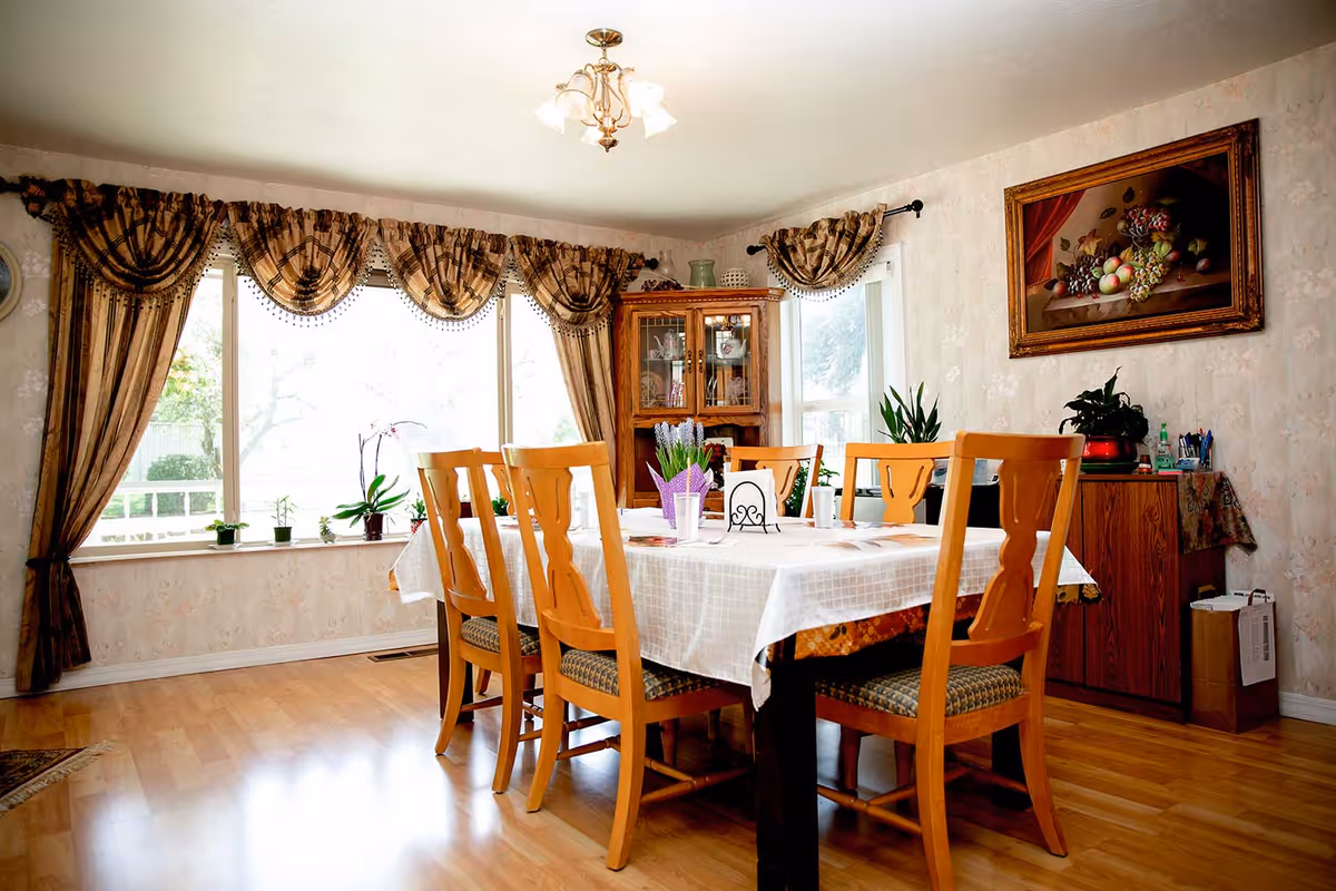 A bright dining room with a wooden table covered by a white tablecloth surrounded by six chairs, large curtained windows, plants, and a china cabinet.