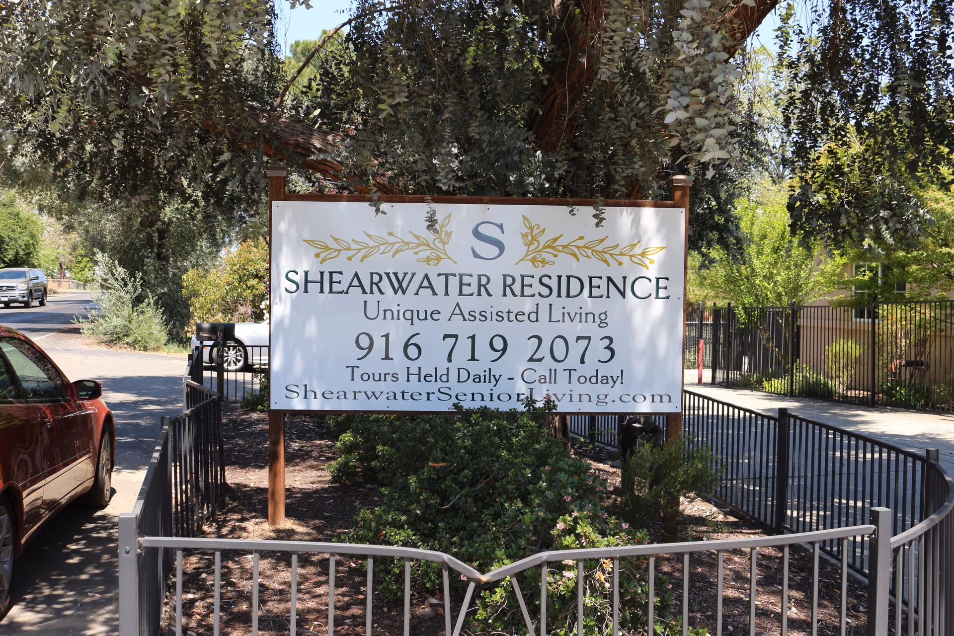 Outdoor view of a sign for Shearwater Residence, an assisted living facility. The sign includes the facility's name, phone number, and website, surrounded by trees, bushes, and a black metal fence. A red car is parked on the left side of the image.