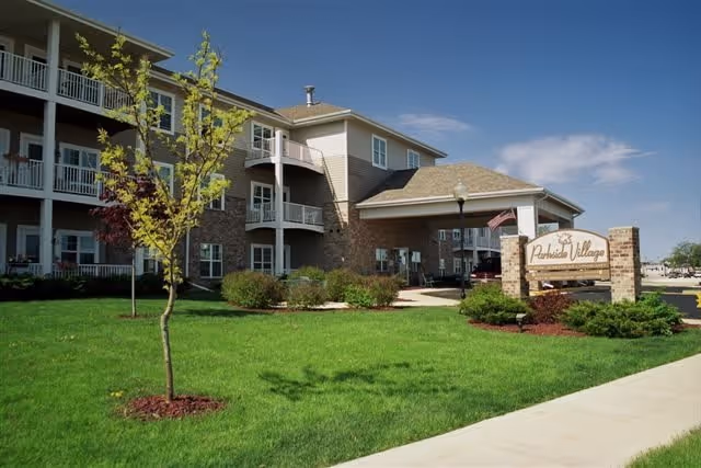 Exterior view of Parkside Village of Hartford, a multi-story senior living facility with balconies, a covered entrance, well-maintained green lawn, small trees, and a sign displaying the facility's name.