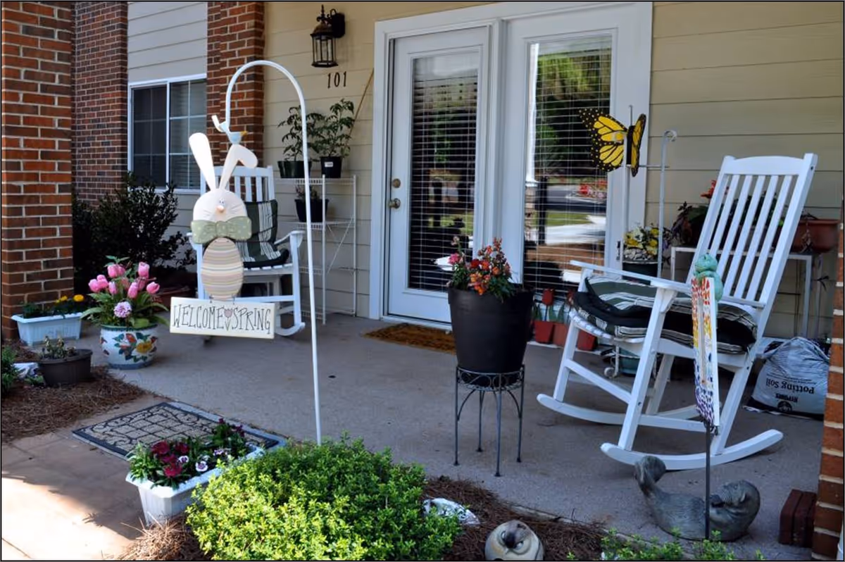 Front porch with rocking chairs, potted plants, and a 'Welcome Spring' bunny sign in front of double glass doors.