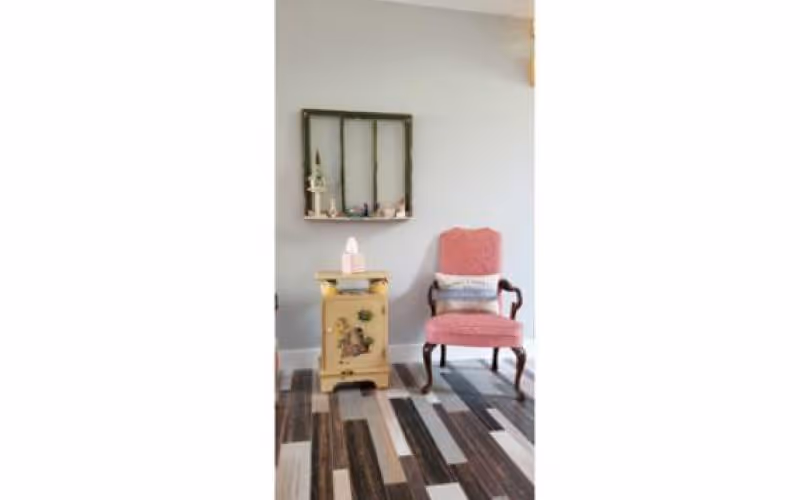 A cozy corner featuring a pink upholstered armchair with a cushion, a small vintage yellow side table with decorative items on top, and a wall-mounted shelf with small decorative objects against a light gray wall. The floor has a patterned wood design in various shades of brown and gray.