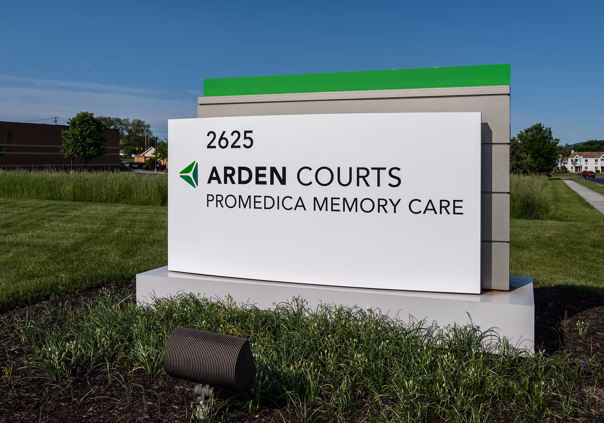 A large white roadside sign reading "2625 ARDEN COURTS PROMEDICA MEMORY CARE" on a grassy lawn in front of the facility.